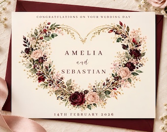 May include: A wedding card featuring a heart-shaped floral wreath of burgundy and blush roses, greenery, and gold accents. The text reads: CONGRATULATIONS ON YOUR WEDDING DAY, AMELIA and SEBASTIAN, 14TH FEBRUARY 2026. The card rests on a dark red envelope.