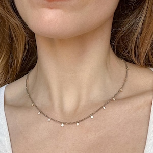 May include: Delicate gold necklace with small, teardrop-shaped pendants evenly spaced along the chain. The necklace is worn around the neck, and the pendants catch the light, creating a subtle sparkle. The necklace is a simple yet elegant piece of jewelry.
