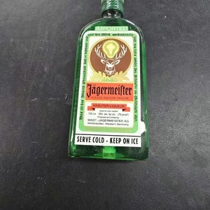 May include: A green glass bottle of Jägermeister herbal liqueur. The label features a stag with a glowing cross between its antlers. The bottle contains 750 ml and is 35% alcohol by volume. The text "SERVE COLD - KEEP ON ICE" is printed on the label.