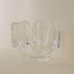 Op de afbeelding: Een heldere glazen kom met een unieke, golvende rand. De kom heeft een brede, ondiepe basis en een reeks verticale, schuine panelen die een textuur en geometrisch uiterlijk creëren. Het transparante glas laat licht door.