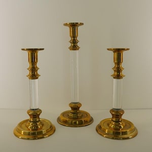 May include: Three decorative candle holders. The bases and tops are gold-coloured, with a clear, cylindrical centre. The candle holders are of varying heights, with the tallest one in the centre.