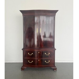 May include: A tall, dark mahogany wardrobe with two doors and two drawers. The wardrobe has gold-colored hardware and ornate carvings along the top. The wood grain is visible, adding to the antique aesthetic.