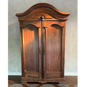 May include: A tall, dark brown wooden armoire featuring two doors with arched panels, a prominent arched cornice top, and decorative carved details. The wood grain is visible across the surface, and the doors include vertical metal handles.