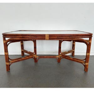 May include: A rectangular coffee table with a clear glass top and a dark brown rattan frame. The frame has curved legs and is reinforced with woven rattan accents. The table is approximately 48 inches long and 24 inches wide.