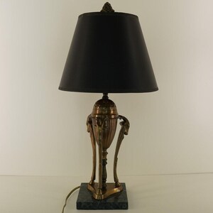 May include: An antique brass table lamp with a black lampshade. The lamp has a decorative urn-shaped body with ornate details and stands on a square green marble base. The lamp is approximately 24 inches tall.