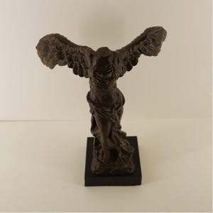 Puede incluir: Estatua de bronce de la Victoria de Samotracia, una figura sin cabeza con alas extendidas. La estatua está montada sobre una base rectangular negra. La escultura representa un drapeado fluido y plumas de ala detalladas.
