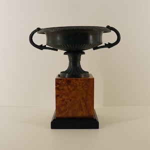 Puede incluir: Una urna de color bronce oscuro con dos asas, colocada sobre un pedestal cuadrado de madera con acabado de madera de raíz. El pedestal descansa sobre una base negra. La urna tiene un diseño acanalado detallado.