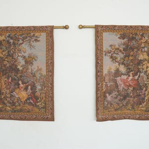 May include: Two rectangular tapestries with a pastoral scene, featuring figures in period clothing, trees, and a building. The tapestries are framed with a brown border and hung from gold-colored rods.