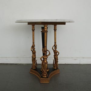 May include: A marble-topped side table with a gold-colored base and three supporting columns. The columns feature classical female figures. The table has a black and gold base and a central black column.
