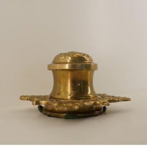 Op de afbeelding: Een vintage messing inktpot met een decoratief bloemmotief op het deksel. De inktpot heeft een brede basis met een sierlijk, stervormig ontwerp en een cilindrisch lichaam. Het stuk is gemaakt van messing en heeft een gepolijste afwerking.