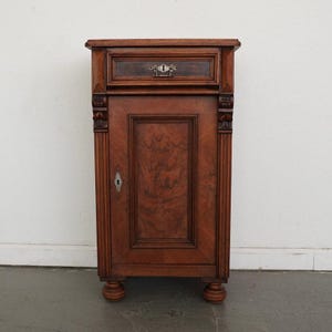 May include: A vintage wooden cabinet with a drawer and door. The cabinet is a rich brown color with intricate wood grain patterns. It features decorative carvings on the sides and a keyhole on the door. The cabinet rests on four rounded feet.