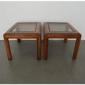 May include: Two square wooden side tables with glass tops. The tables are a medium brown color and have a simple, classic design. Each table has four legs and a glass insert in the top. The tables are approximately 24 inches wide and 24 inches deep.