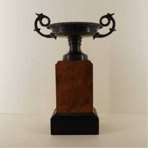 Puede incluir: Un trofeo decorativo con un cuenco y asas de bronce oscuro, colocado sobre un bloque de madera marrón y una base negra. El cuenco tiene un diseño de borde detallado. El trofeo es un diseño clásico.