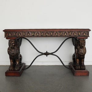 May include: A dark wood console table with ornate detailing. The table features two bronze-colored lion sculptures with wings as supports. The tabletop has a decorative edge with gold accents, and the base is supported by a black metal frame.