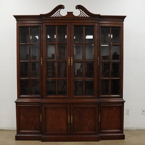 May include: A large, dark wood cabinet with glass-paned doors and brass hardware. The cabinet has a decorative top with a carved design and a base with solid doors. The interior is visible through the glass.