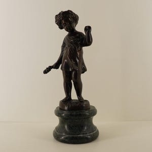 Puede incluir: Una escultura de bronce de un niño pequeño sosteniendo un objeto floral, de pie sobre un pedestal de mármol verde oscuro. El niño lleva una prenda drapeada. La escultura es una obra de arte decorativa.