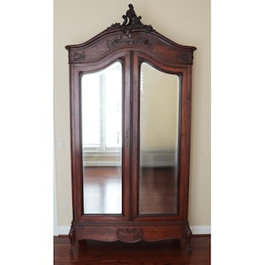 May include: A tall, dark brown wooden armoire featuring two full-length mirrored doors with arched tops. The piece has an ornate, carved wooden crest at the top center and stands on four curved legs, with a carved decorative panel at the base.