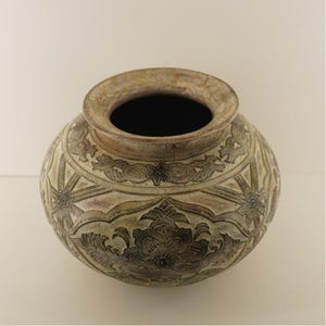 May include: A round, beige ceramic pot with a wide opening and intricate floral and geometric relief patterns. The pot's surface has a weathered appearance, with darker shading in the recessed areas, enhancing the details of the design.