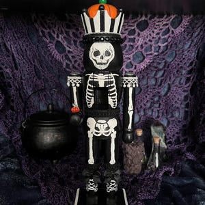 May include: A black and white skeleton nutcracker with a pumpkin crown and a skull face. The Halloween-themed decoration has a black base and is adorned with bows and a small pumpkin on its arm. The background includes a cauldron and bottles.