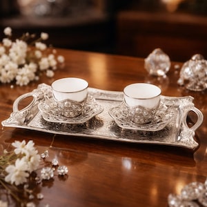 Puede incluir: Una bandeja plateada con dos tazas de té blancas y sus platillos. La bandeja y los platillos tienen diseños ornamentados en relieve. Las tazas tienen un borde plateado. La escena está ambientada en una mesa de madera oscura, con flores y cristales decorativos.