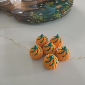 Op de afbeelding: Zes kleine, oranje pompoenversieringen met groene stelen en groene gesneden lijnen. De pompoenen zijn gerangschikt op een wit oppervlak, met een decoratief houten dienblad op de achtergrond.