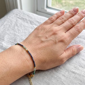 May include: A delicate beaded bracelet featuring small, faceted dark blue and light green gemstone beads arranged in a gradient pattern. The bracelet includes tiny gold-toned metal spacer beads and a gold-toned metal lobster clasp with an extender chain.