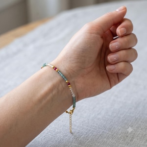 May include: A delicate beaded bracelet featuring small, faceted blue-green and clear beads interspersed with vibrant pink round beads and tiny gold-toned metallic spacers. The bracelet includes a gold-toned metal chain extender with a lobster clasp.