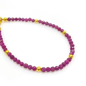 May include: A delicate bracelet featuring small, faceted, deep pink gemstones strung together. The bracelet is accented with gold-colored beads and a gold-colored clasp. The jewelry is displayed on a white background.
