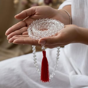 Puede incluir: Un collar de mala de cristal de cuarzo transparente con una borla roja. Las cuentas son redondas y están ensartadas, formando una hebra larga. El collar se sostiene en manos abiertas, sobre un fondo blanco.