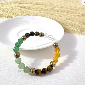 May include: A multi-gemstone bracelet with green, gold, brown, and clear beads. The bracelet includes green aventurine, pyrite, tiger's eye, and clear quartz beads. The bracelet is displayed on a white plate. The beads are approximately 0.8 cm in diameter.