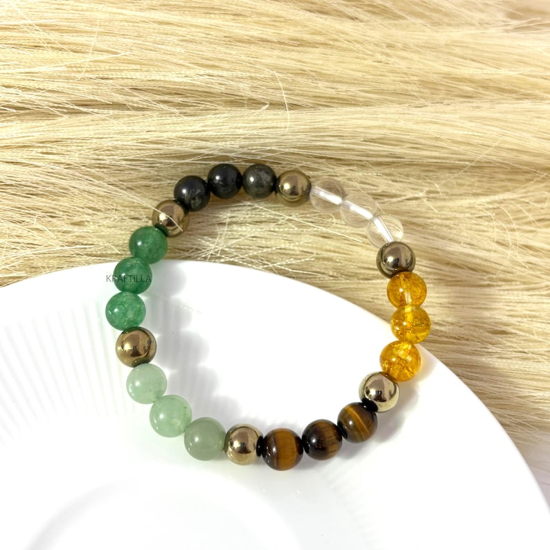 May include: A multi-gemstone bracelet with green, gold, brown, and clear beads. The bracelet is displayed on a white plate, set against a neutral backdrop. The beads are spherical and of varying sizes.
