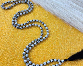 May include: A beaded necklace with white and black speckled beads. The necklace is draped on a yellow textured surface, with a white feathery accent in the background. The necklace has a black tassel.