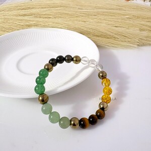 May include: A multi-gemstone bracelet with green, gold, brown, and clear beads. The bracelet includes green aventurine, pyrite, tiger's eye, and clear quartz. The bracelet is displayed on a white plate, with dried ornamental grass in the background.