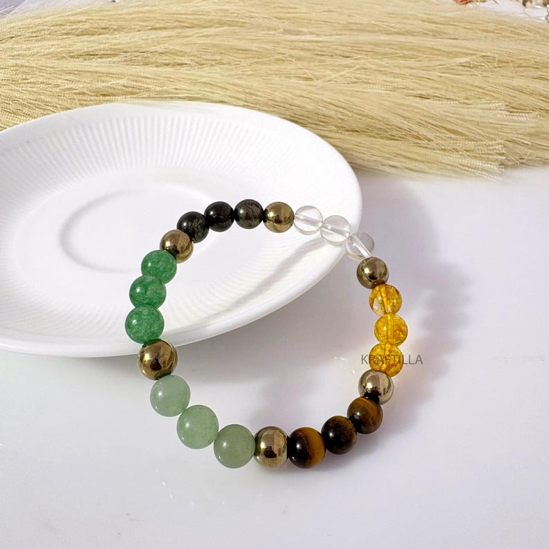 May include: A multi-gemstone bracelet with green, gold, brown, and clear beads. The bracelet includes green aventurine, pyrite, tiger's eye, and clear quartz. The bracelet is displayed on a white plate, with dried ornamental grass in the background.