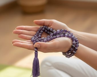 Op de afbeelding: Een paarse kralen mala ketting met een bijpassende kwast. De mala wordt in open handen gehouden, met de nadruk op de gladde, ronde kralen. De achtergrond is wazig, waardoor de ketting wordt benadrukt.