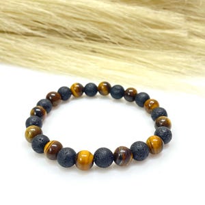 Könnte beinhalten: Ein Armband mit abwechselnd schwarzen Lavastein- und Tigeraugenperlen. Die Tigeraugenperlen zeigen eine Mischung aus Braun-, Gold- und Gelbtönen. Das Armband wird auf einer weißen Oberfläche mit einem verschwommenen beigen Hintergrund präsentiert.