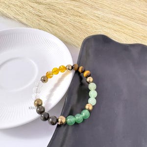 May include: A gemstone bracelet with clear, yellow, brown, gold, and green beads. The bracelet is presented on a white plate and a black tray. The beads are likely around 0.8 cm in diameter.