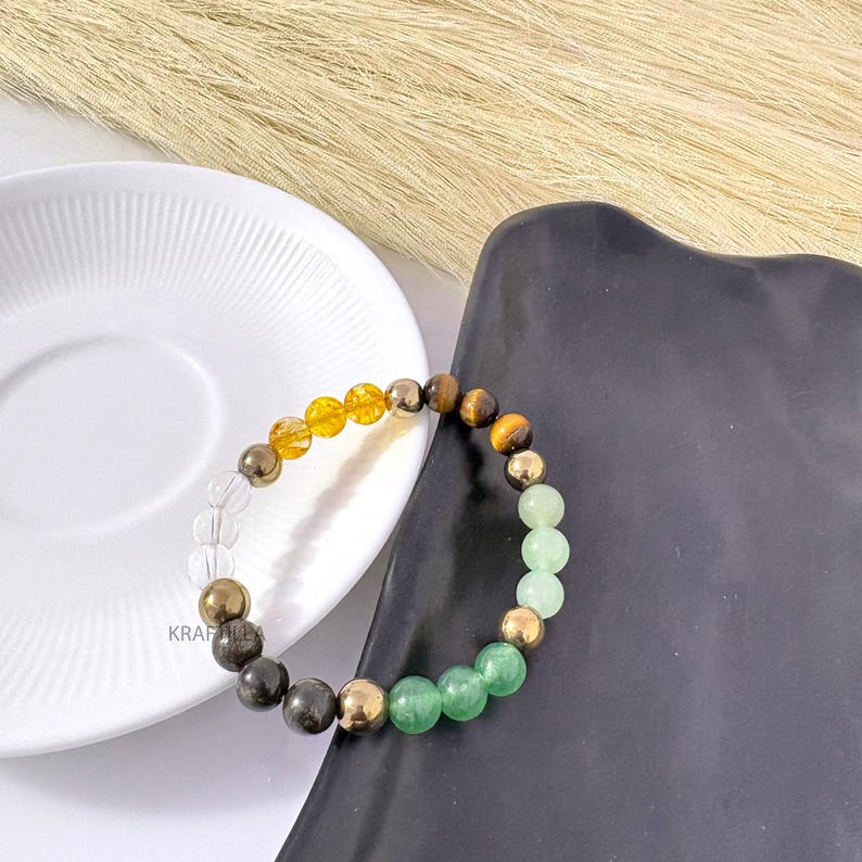 May include: A gemstone bracelet with clear, yellow, brown, gold, and green beads. The bracelet is presented on a white plate and a black tray. The beads are likely around 0.8 cm in diameter.