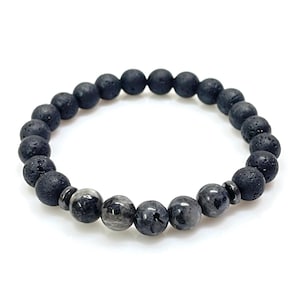 Può includere: Un braccialetto di perline nere con una miscela di perline di roccia lavica strutturate e perline di pietre preziose grigie lisce. Il braccialetto è infilato su un elastico, mostrando un design semplice e minimalista. Le perline hanno un diametro di circa 0,8 cm.