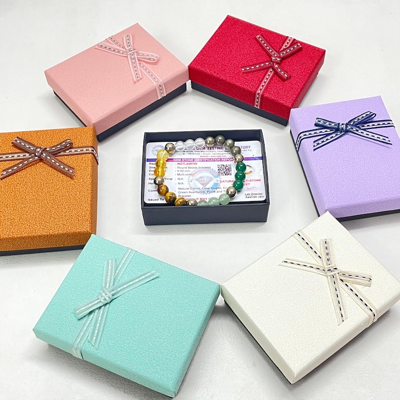May include: A gemstone bracelet with yellow, brown, green, and silver beads is displayed on a card within a black box. The box is surrounded by six colourful gift boxes in pink, red, orange, purple, white, and teal, each tied with a ribbon.