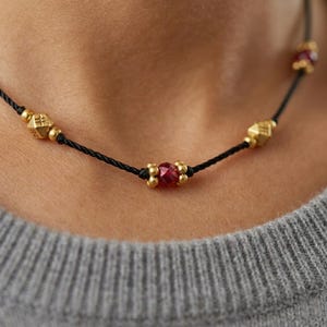 May include: A close-up of a necklace with a black cord. The necklace features gold-colored beads and a central red gemstone. The gold beads have a textured design. The necklace is worn against a gray sweater.