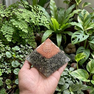 May include: A pyramid-shaped resin object featuring a clear base filled with dark metallic pyrite-like mineral fragments, topped with a layer of copper-colored shavings and a small, clear, stacked-disc crystal point at the very peak.
