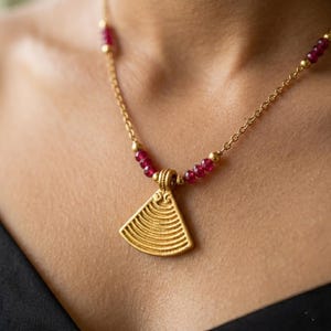 Puede incluir: Un collar dorado con un colgante en forma de abanico triangular. El collar presenta pequeñas cuentas rojas redondas y una delicada cadena. El colgante tiene un diseño texturizado y acanalado.