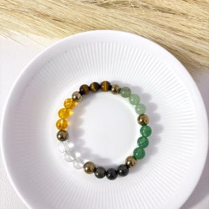 May include: A multi-coloured gemstone bracelet. The bracelet features clear, yellow, brown, green, and gold beads. The bracelet is displayed on a white plate with a ridged design.