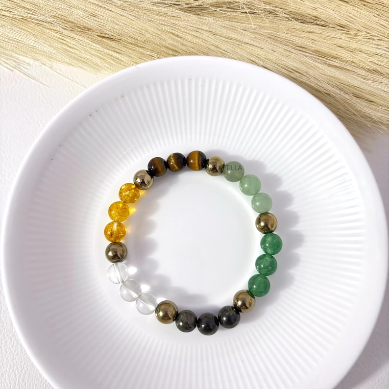 May include: A multi-coloured gemstone bracelet. The bracelet features clear, yellow, brown, green, and gold beads. The bracelet is displayed on a white plate with a ridged design.