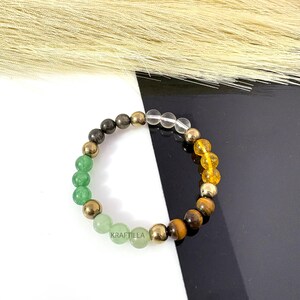 May include: A bracelet with a mix of gemstones and gold-coloured beads. The bracelet features green, clear, yellow, and brown beads. The word "KRAFTILLA" is printed on the bracelet.
