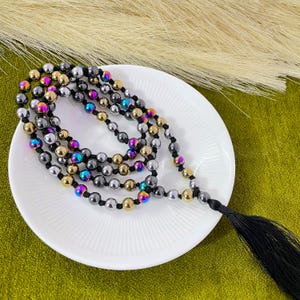 May include: A beaded necklace with a black tassel. The necklace features a mix of metallic beads in gold, silver, and iridescent colors. The necklace is draped on a white plate with a green background.