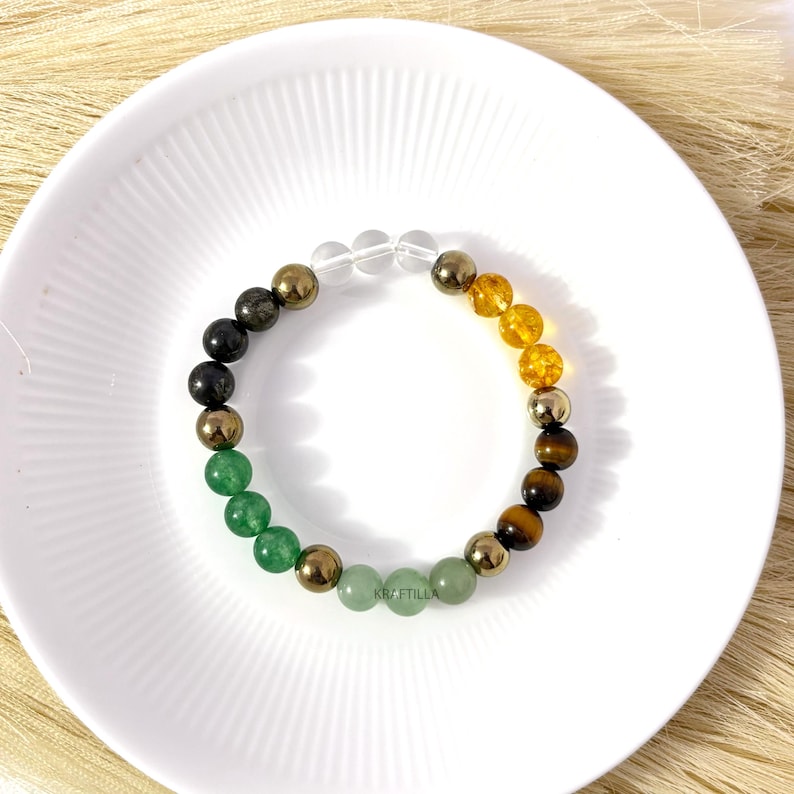 May include: A colourful beaded bracelet with green, gold, black, clear, and amber-coloured gemstones. The bracelet is displayed on a white plate. The beads are of varying sizes.
