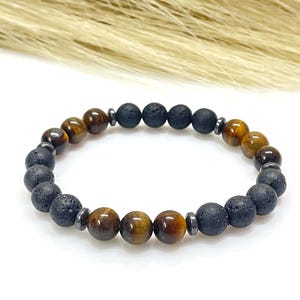 Puede incluir: Una pulsera de cuentas con ojo de tigre, lava negra y cuentas espaciadoras plateadas. Las cuentas de ojo de tigre muestran tonos marrones y dorados cálidos, mientras que las cuentas de lava tienen una textura porosa. La pulsera está ensartada en un cordón elástico.