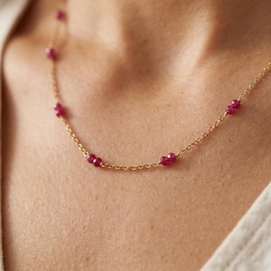 May include: A delicate gold necklace featuring small, vibrant red gemstones. The necklace is composed of a fine gold chain with evenly spaced, round ruby beads. The necklace is shown against a neutral background, highlighting the jewelry's simple elegance.